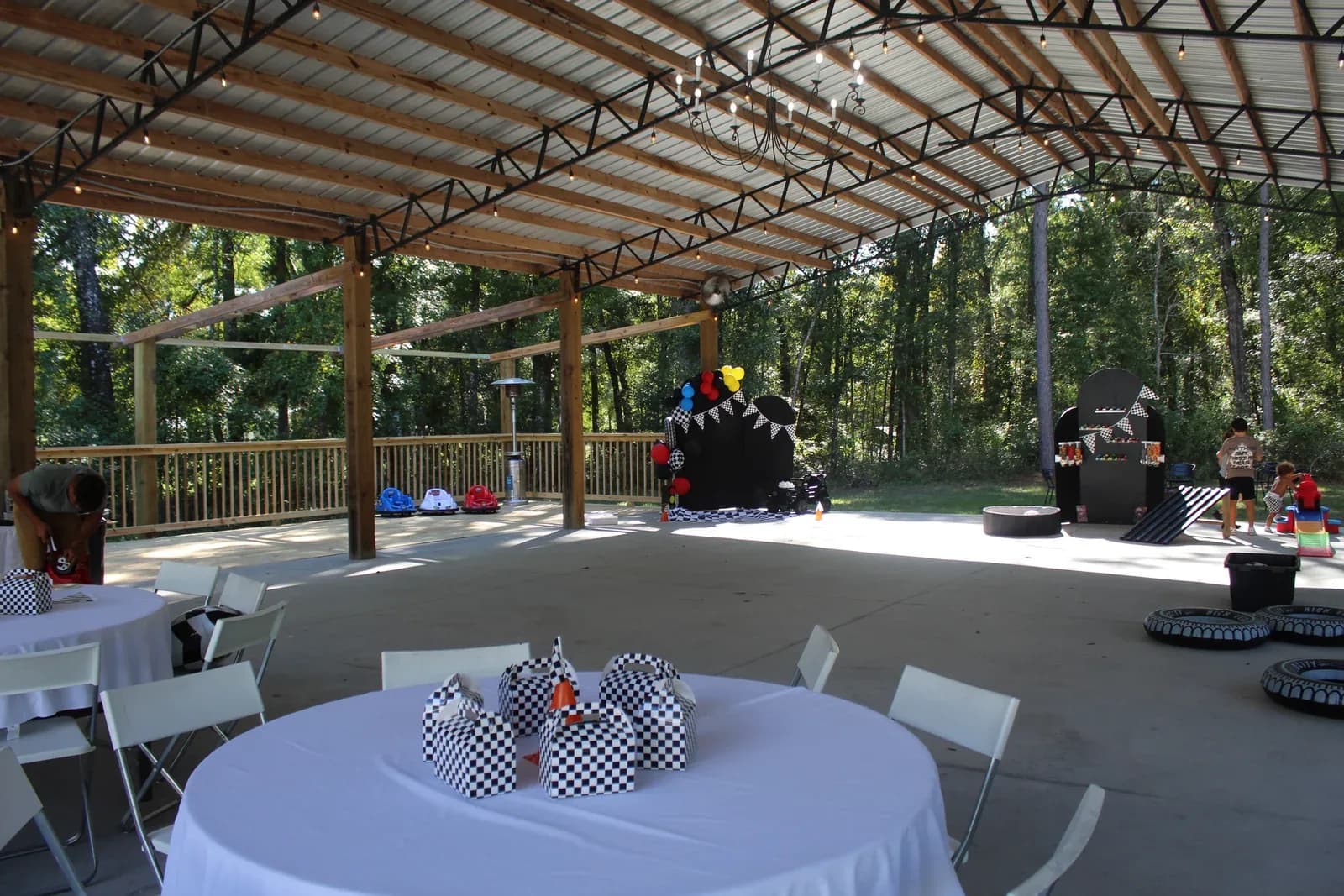 A children's birthday party set up under the pavilion, white tablecloths with checkered favor boxes, a race car backdrop with balloons and ride-on cars.