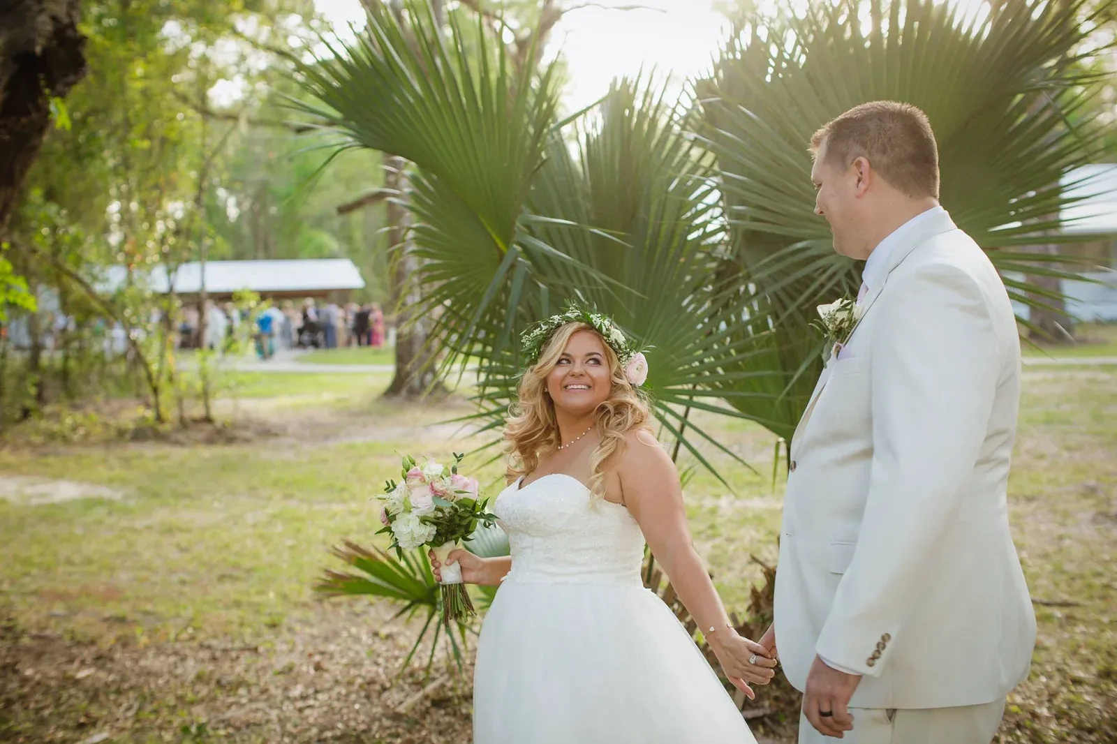 A bride in a flower crown, holding a pink and white bouquet, smiles back at her groom in a cream suit as they hold hands beside a stand of palms.