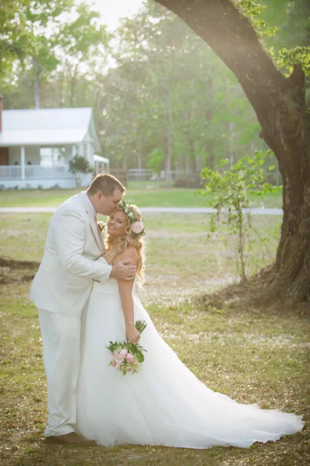 A groom in a cream suit kisses his bride on the forehead beneath a large oak, her train spread across the grass and the farmhouse behind them.