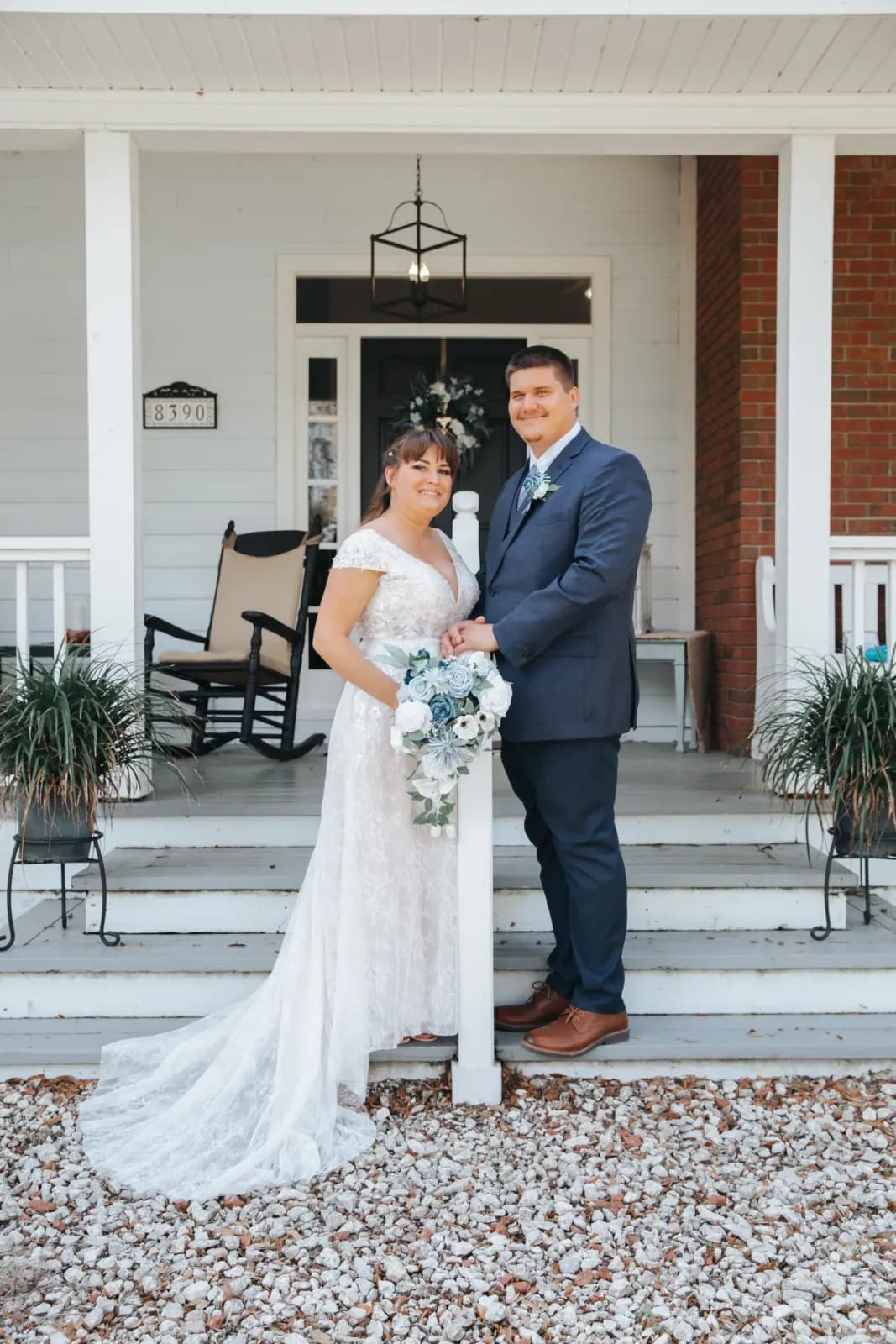 A bride and groom standing together at the foot of the farmhouse steps, a rocking chair and potted plants on the porch behind them.