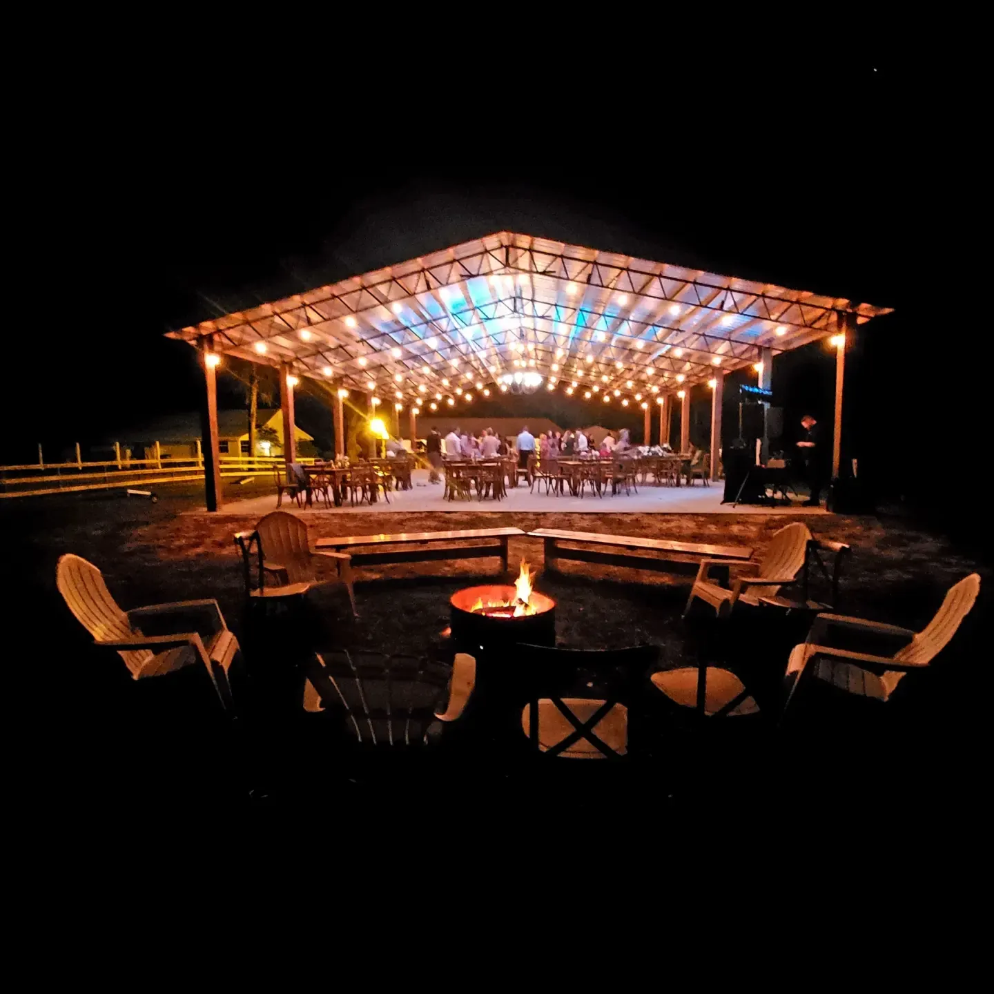 The open barn lit up after dark, string lights and stage lighting along the trusses with guests seated at tables inside, and a ring of wooden chairs around a lit fire pit in the foreground.