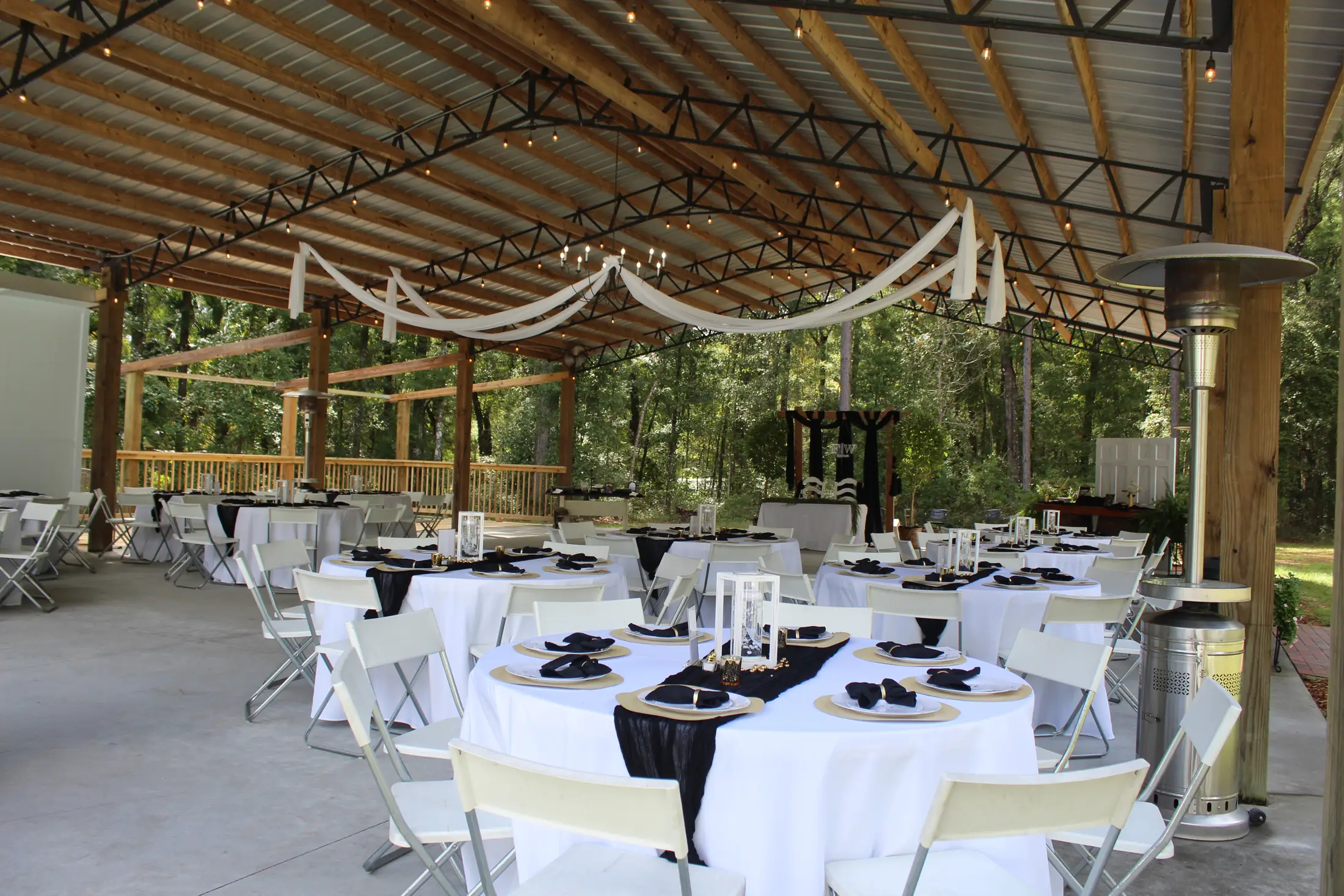 The open sided barn set for a reception: round tables in white cloths with black runners and gold chargers, folding chairs, white fabric swagged along the steel trusses, string lights overhead and woodland visible past the posts.
